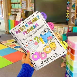 May include: A spiral bound notebook with a colorful cover featuring the text "Daily 5 Yearly Program Weekly Overviews" and "Grades 1 & 2". The cover also has illustrations of letters, a pencil, and a book with googly eyes.