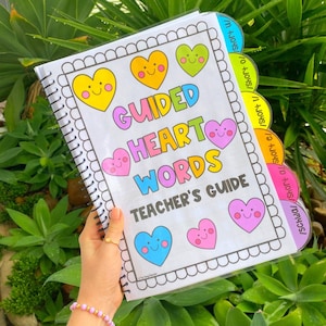 May include: A spiral bound teacher's guide for guided heart words. The cover features colourful hearts with smiling faces and the title "Guided Heart Words Teacher's Guide". The guide has tabs labelled with short vowel sounds.