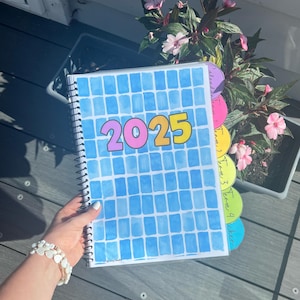 May include: A blue and white planner with a watercolour design of a swimming pool. The planner has tabs for each month, labelled "Term 1", "Term 2", "Term 3", and "Term 4". The front cover of the planner has the year "2025" written in large, colourful letters.
