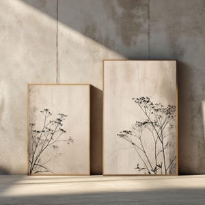 Könnte beinhalten: Zwei gerahmte botanische Drucke. Jeder Druck zeigt einen beigefarbenen Hintergrund mit einem schwarz-weißen Blumendesign. Die Rahmen sind hellbraun. Die Drucke werden vor einer strukturierten, hellgrauen Wand ausgestellt.