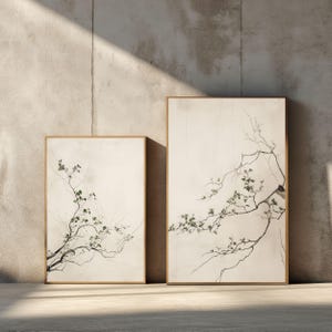 May include: Two rectangular wall art prints in thin wooden frames. Each print features a cream-colored background with a delicate, dark brown tree branch extending across the frame, adorned with small, sparse green leaves and thin twigs.