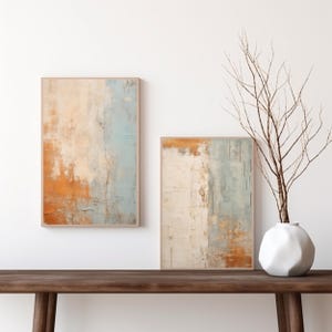 Puede incluir: Dos pinturas abstractas en marcos de madera, una encima de la otra, con una gama de colores marrón claro, blanco y azul. Las pinturas están colgadas en una pared blanca sobre una mesa de madera marrón oscuro con un jarrón blanco y una rama con ramas desnudas.