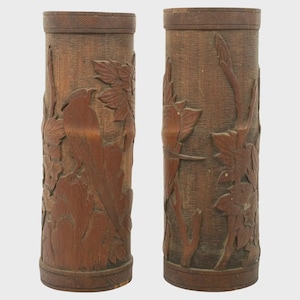 Peut inclure: Deux vases cylindriques en bois avec des sculptures en relief détaillées. Les vases sont de couleur marron chaud et présentent des représentations d'oiseaux, de feuilles et d'éléments floraux. Les bords supérieurs et inférieurs ont un motif géométrique décoratif.