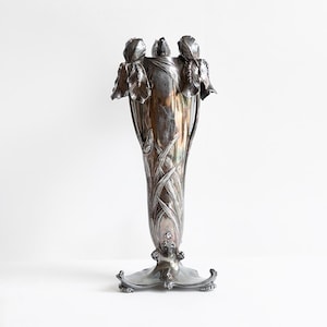 Peut inclure: Un grand vase en métal argenté doté d'un corps fuselé orné de motifs de fleurs d'iris en relief sur le bord et de longues feuilles fines descendant le long des parois. La base est large et travaillée avec des pieds décoratifs incurvés.