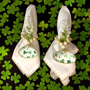 Puede incluir: Dos servilletas de lino blanco dobladas, adornadas con servilleteros con temática del Día de San Patricio. Cada servilletero presenta un dije en forma de concha con un patrón de trébol verde y ribete dorado. Guirnalda de cuentas y vegetación artificial.