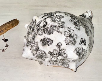 Könnte beinhalten: Eine weiße Muschelschale mit einem schwarzen Blumen- und Schmetterlingsdesign. Die Schale hat eine strukturierte Oberfläche und eine natürliche, organische Form. Der Schwarz-Weiß-Kontrast hebt die komplizierten Details des Kunstwerks hervor.