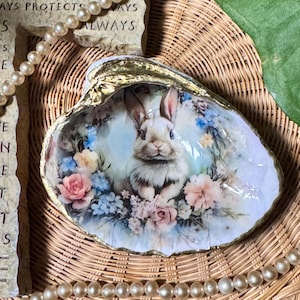 Puede incluir: Concha decorativa con una imagen pintada de un conejo blanco rodeado de flores rosas y azules. La concha tiene un borde dorado y descansa sobre una superficie de mimbre. Un collar de perlas y una piedra con la palabra "ALWAYS" están en el fondo.