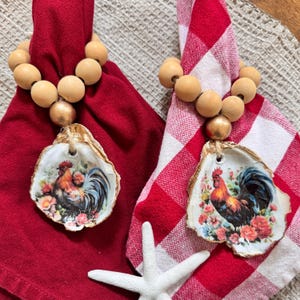 May include: Two napkin rings with wooden beads and oyster shell pendants. Each shell features a rooster illustration with floral accents. One napkin is red, the other is red and white checkered.