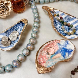 Puede incluir: Tres platos decorativos de concha de ostra con ribete dorado. Un plato tiene un patrón floral azul y blanco, otro tiene un patrón floral rosa y azul con una estrella de mar azul, y el tercero tiene un patrón a rayas azul y blanco. Los platos están sobre una superficie blanca con una hebra de cuentas azules y doradas.
