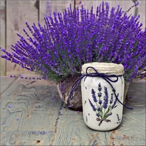Peut inclure: Un pot blanc décoré de lavande, noué avec une ficelle bleu marine, posé sur une surface en bois vieilli. Un pot de fleurs de lavande violettes est en arrière-plan, créant un thème floral rustique.