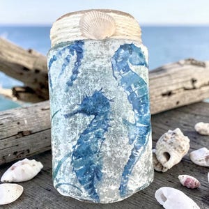 May include: A cylindrical glass jar with a blue seahorse design. The jar is decorated with a rope and a seashell on the top. The jar is sitting on a wooden surface with seashells scattered around it.