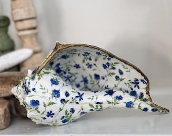 Könnte beinhalten: Eine weiße Muschel mit einem blauen und grünen Blumenmuster. Die Muschel ist mit goldenen Akzenten bemalt und hat eine glatte, glänzende Oberfläche.