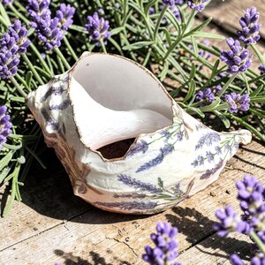 Könnte beinhalten: Eine dekorative Muschelschale mit weißem Innenraum und Außenseite, verziert mit Lavendelzweigen. Der Rand hat eine goldene Zierleiste. Die Schale steht auf einer Holzoberfläche, umgeben von frischen Lavendelzweigen mit lila Blüten.