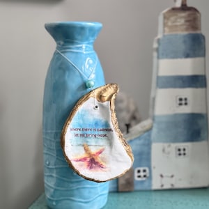 Puede incluir: Un jarrón de cerámica azul con una concha de ostra decorativa. La concha tiene un borde dorado, una imagen de acuarela de una estrella de mar y cielo, y el texto "Where there is sadness, let me bring hope."