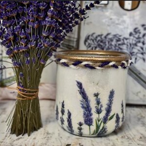 Peut inclure: Un pot en verre blanc avec un bord doré et un détail de corde tressée, décoré de brins de lavande violets. Un bouquet de lavande séchée est attaché avec de la ficelle à côté du pot. L'arrière-plan comprend une impression sur le thème de la lavande.