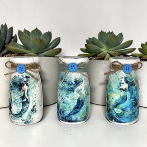 Puede incluir: Tres tarros de vidrio decorativos con diseños de sirenas. Cada tarro presenta una tapa blanca, un botón azul y detalles de cuerda. El arte de la sirena está en tonos azules y verdes. Las suculentas están en el fondo.
