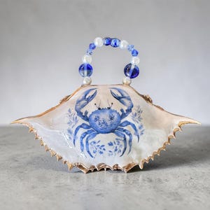 May include: A decorative crab shell ornament featuring a hand-painted blue crab illustration with floral accents. The shell has a gold-painted serrated edge and a handle constructed from alternating white pearl-like beads and translucent blue glass beads.