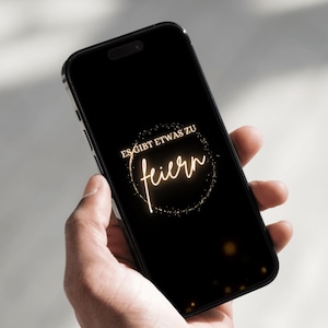 Puede incluir: Un smartphone negro muestra un mensaje de celebraci&oacute;n dorado sobre un fondo negro. El texto dice "ES GIBT ETWAS ZU feiern" en un dise&ntilde;o circular, sugiriendo una ocasi&oacute;n festiva.