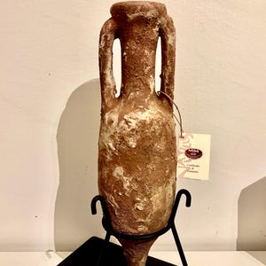 Può includere: Un'anfora in terracotta con due manici, una base a punta e una superficie ruvida e testurizzata. L'anfora è su un supporto in metallo nero e ha un'etichetta bianca con il testo "N.B.S. Certificato di Garanzia".