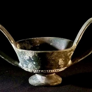 Peut inclure: Une coupe en bronze avec deux anses et une base circulaire. La coupe a un aspect usé et vieilli et une bande décorative autour du bord.