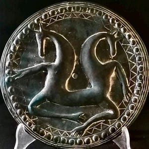Pode incluir: Um espelho circular de metal cinza escuro com um design em relevo de duas criaturas míticas. O espelho é rodeado por uma borda decorativa de círculos e triângulos.