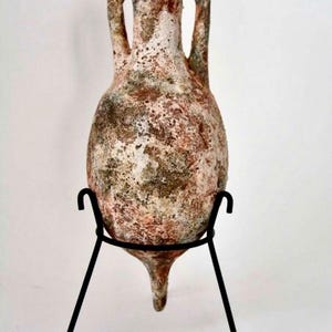 Può includere: Un'anfora in terracotta con una smaltatura bianca e marrone screziata, su un supporto in metallo nero. L'anfora ha due manici e una base a punta.