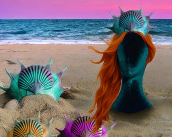 May include: A teal and purple seashell crown with orange hair on a mannequin head, and three seashell crowns on a sandy beach with the ocean and a pink and blue sky in the background. The crowns have a spiky design.