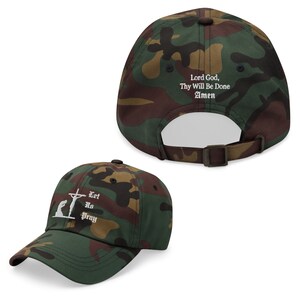 May include: Camouflage baseball cap with white text and a religious theme. The front of the cap features an image of a person kneeling before a cross with the words "Let Us Pray." The back of the cap has the words "Lord God, Thy Will Be Done Amen."