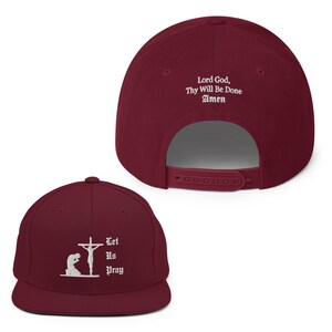 May include: Maroon flat-brimmed baseball cap with white embroidered religious imagery. The front features a kneeling figure before a cross with the words "Let Us Pray." The back displays "Lord God, Thy Will Be Done Amen."