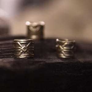Könnte beinhalten: Drei goldfarbene Haarperlen aus Metall mit aufwendigen geometrischen Mustern. Die Perlen sind zylindrisch und liegen auf einer dunklen, strukturierten Oberfläche. Die Beleuchtung erzeugt ein weiches, diffuses Leuchten.