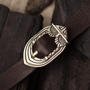 May include: A brown leather belt with a decorative silver-colored buckle. The buckle features an owl design and is set against a backdrop of dark wood and charcoal.