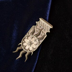 Op de afbeelding: Zilverkleurige metalen broche met een bloemmotief in het midden, omringd door gestileerde vlammen. De rechthoekige broche heeft een decoratieve bovenrand. Het ligt op een donkerblauwe en zwarte fluwelen achtergrond.