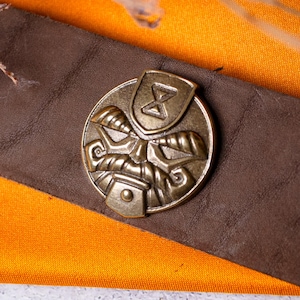 Op de afbeelding: Een bruine leren riem met een ronde koperen gesp met een reliëfontwerp van een gestileerd gezicht en een zandlopersymbool aan de bovenkant. De gesp heeft een metalen afwerking en is gecentreerd op de brede, getextureerde bruine leren riem.