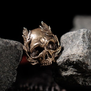 Puede incluir: Un anillo de calavera de oro con un diseño de corona de laurel. El anillo está colocado sobre un fondo de rocas grises oscuras y una cadena.