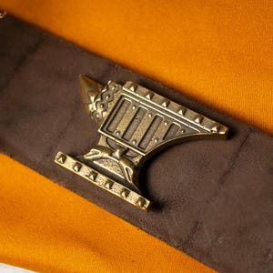 May include: A metallic brass-toned belt buckle shaped like an anvil, featuring a textured surface with small pyramid-shaped studs along the base and top edges, attached to a wide, dark brown leather belt strap with a smooth, matte finish.