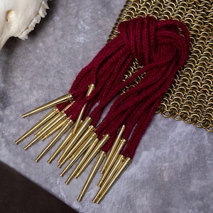 Puede incluir: Un conjunto de cordones de color burdeos con puntas de metal dorado. Los cordones están dispuestos de forma ordenada, con las puntas orientadas hacia afuera. Una sección de cota de malla dorada es visible en el fondo.