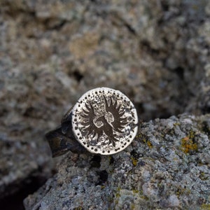 May include: A silver ring with a circular design featuring a stylized eagle with wings spread. The ring is on a rough, gray, textured surface.