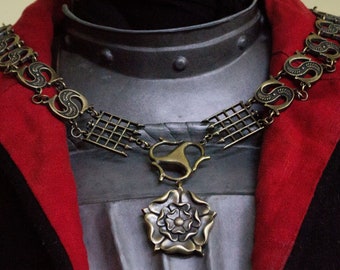 May include: A gold chain necklace with a pendant featuring a stylized rose. The chain is made of interlocking links and has a decorative design. The pendant is a large, ornate rose with a central pearl.