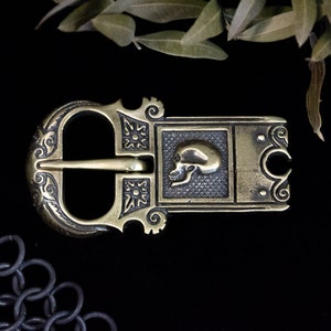 Puede incluir: Una hebilla de cinturón de latón con un diseño ornamentado que presenta una calavera en el centro. La hebilla es de color dorado oscuro y tiene un diseño detallado e intrincado.