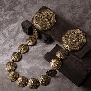 Puede incluir: Un collar dorado y dos colgantes octogonales a juego con intrincados diseños en relieve. El collar presenta eslabones redondos en forma de escudo. Las joyas se exhiben sobre bloques de madera oscura sobre un fondo gris moteado.