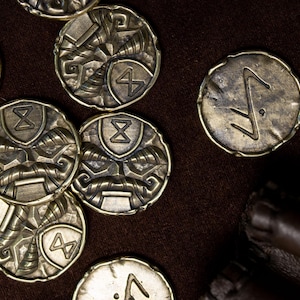 Peut inclure: Une collection de pièces de monnaie métalliques circulaires couleur laiton présentant des motifs en relief de visages stylisés avec moustaches et des symboles géométriques semblables à des runes. Finition antique patinée et sombre.