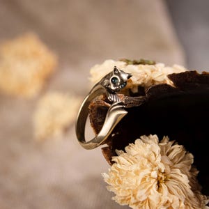Könnte beinhalten: Ein goldfarbener Katzenring mit blauen Edelsteinaugen. Der Ring ist um eine getrocknete Blume und ein dunkelbraunes, strukturiertes Objekt gewickelt. Der Hintergrund ist eine weiche, neutrale Farbe.