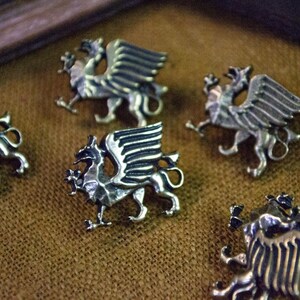 May include: Five silver-toned metal buttons, each shaped like a winged griffin in profile. The buttons feature detailed engravings on the wings and body, with a metallic finish that reflects light, showcasing the mythical creature's form.