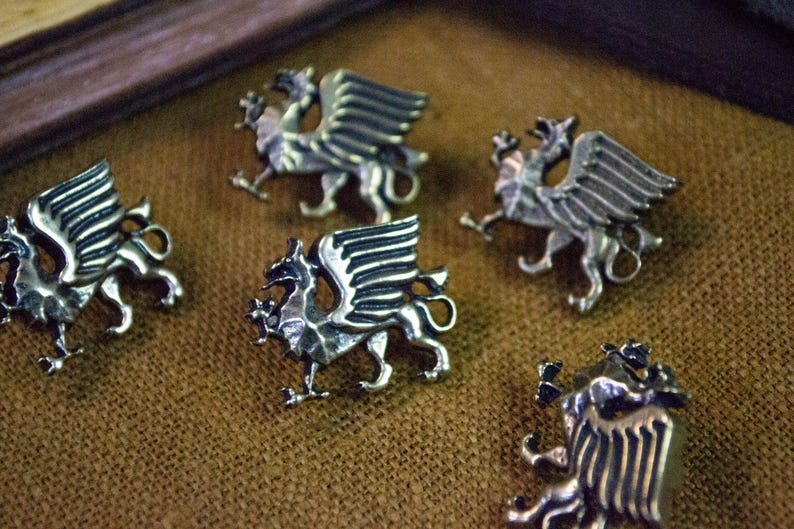 May include: Five silver-toned metal buttons, each shaped like a winged griffin in profile. The buttons feature detailed engravings on the wings and body, with a metallic finish that reflects light, showcasing the mythical creature's form.