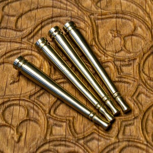 May include: Four gold-colored metal rods with pointed ends, arranged in a fan shape on a wooden surface with a carved pattern.