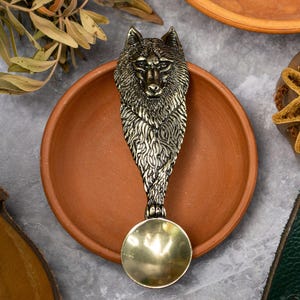 May include: A decorative spoon with a wolf head design. The spoon is made of silver-colored metal and has a round bowl. It rests on a terracotta-colored plate, with other rustic items in the background.