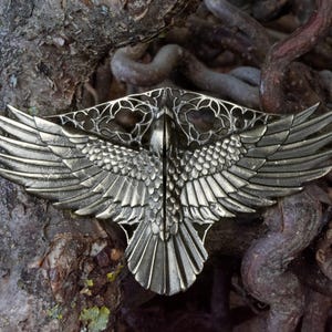Pode incluir: Uma peça decorativa de metal prateado em forma de pássaro com as asas abertas. As asas apresentam padrões de penas detalhados. A peça está colocada sobre um fundo de raízes e galhos de árvores, criando um contraste natural.