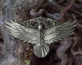 May include: A silver-colored metal decorative piece in the shape of a bird with outstretched wings. The wings have detailed feather patterns. The piece is set against a backdrop of tree roots and branches, creating a natural contrast.