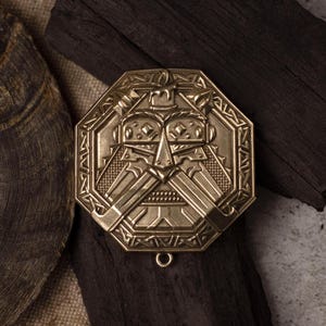 May include: A gold-coloured, octagonal pendant with an engraved face design. The pendant has a loop at the bottom and is set against a backdrop of dark wood and a textured surface. The design features a stylized face with geometric shapes.