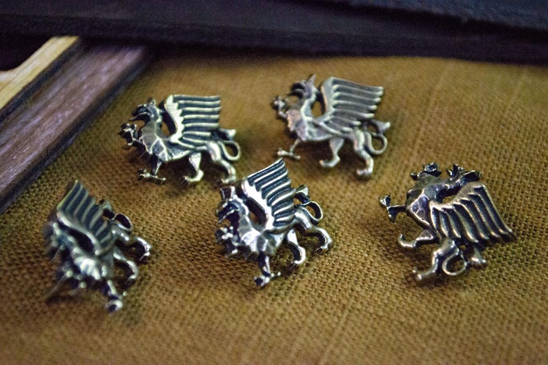 May include: Five silver-toned metal buttons, each shaped like a griffin with wings spread, a curved tail, and a detailed head. The buttons feature a polished metallic finish with dark recessed areas that highlight the sculpted creature design.